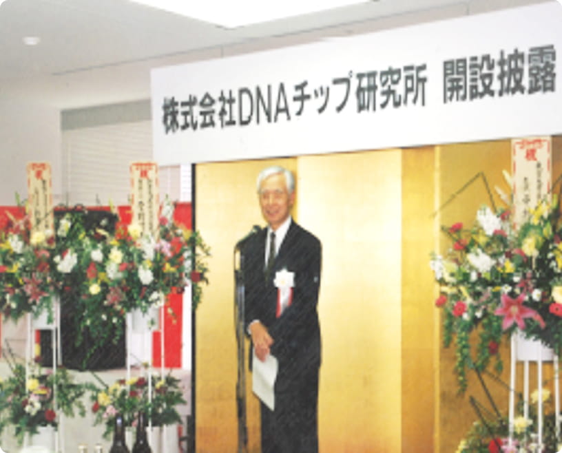 「株式会社DNAチップ研究所 開設披露」と書かれた看板の前でスピーチする男性